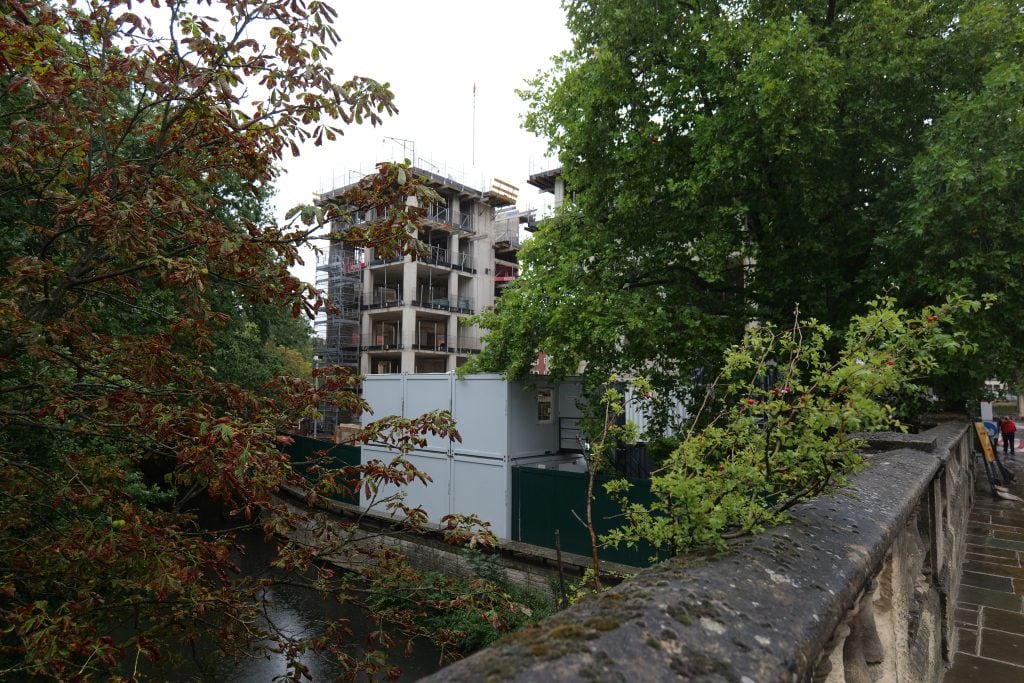 A view from Magdalen Bridge looking towards the Waynflete Quad construction site. The concrete frame and scaffolding of a new building rise behind lush trees, temporary site cabins, and green hoardings along the riverbank.