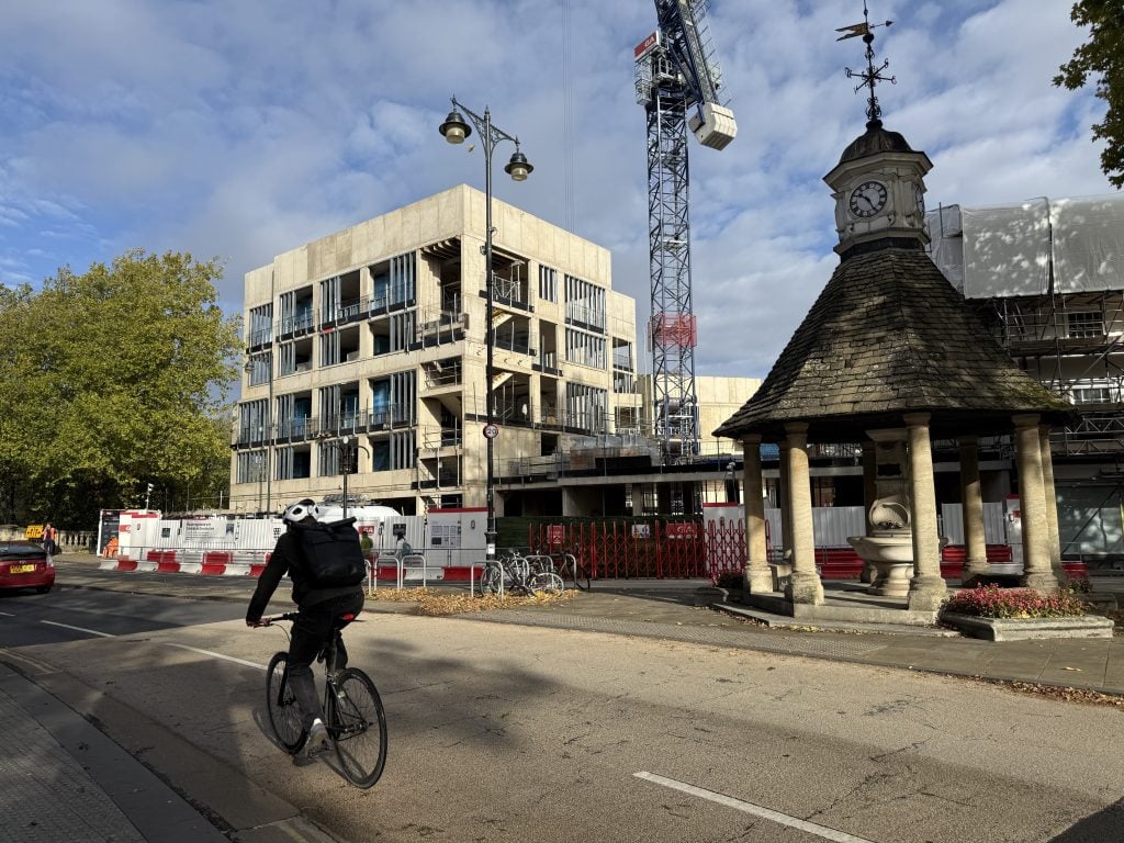 A cyclist rides along the road at The Plain in Oxford, passing the historic clock-faced monument known as the Isis Fountain. In the background, the Waynflete Building at Magdalen College undergoes redevelopment alongside a towering construction crane under a partly cloudy sky.