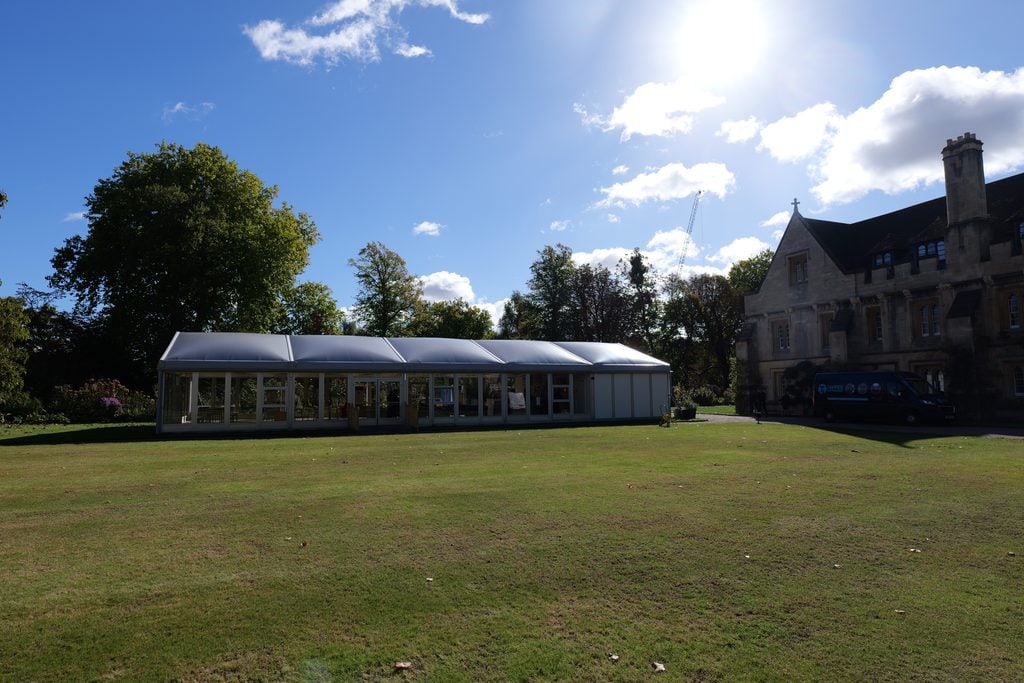 A large white marquee with clear glass windows sits on New Buildings lawn. Cloisters stands on the right under a bright, sunny blue sky with scattered clouds. Lush green trees border the background.
