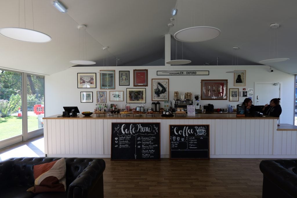New Temporary College Bar: interior with a wooden serving counter and white panelling. Two chalkboard menus displaying food and coffee prices sit on the wooden floor in front of the bar. Framed artwork hangs on the back wall, along with a vintage "Magdalen - Oxford" train station sign. Circular disc pendant lights hang from the high ceiling, while large glass windows show outdoor trees and greenery on the left. Dark leather tufted armchairs rest in the foreground.