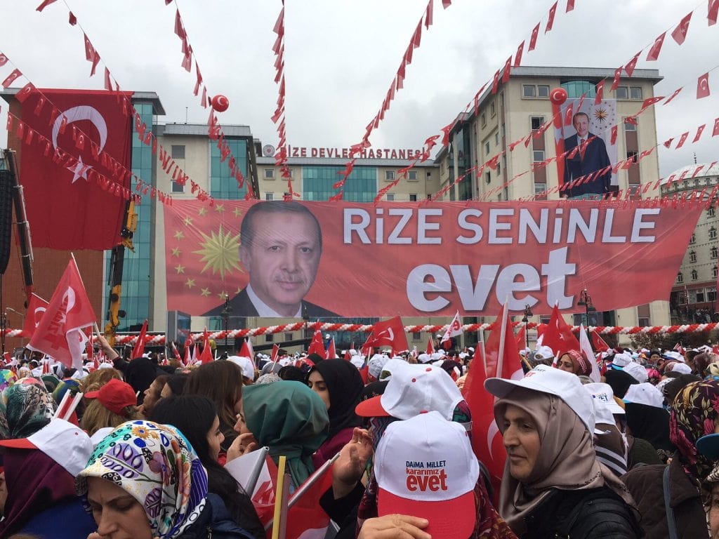 A large crowd gathers outdoors at a political rally in Turkey, holding small Turkish flags. Strings of Turkish bunting stretch across the sky overhead. In the background stands a building with the Turkish inscription "RİZE DEVLET HASTANESİ". A wide red banner displays a portrait of Recep Tayyip Erdoğan beside the text "RİZE SENİNLE evet". Attendees in the foreground wear white hats that feature the word "evet".